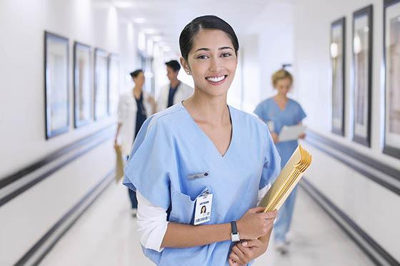 A nurse in blue scrubs holds patient files and smiles for a photo
